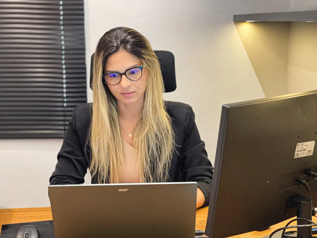 Integrante do escritório de blazer preto e óculos concentrada em notebook e monitor