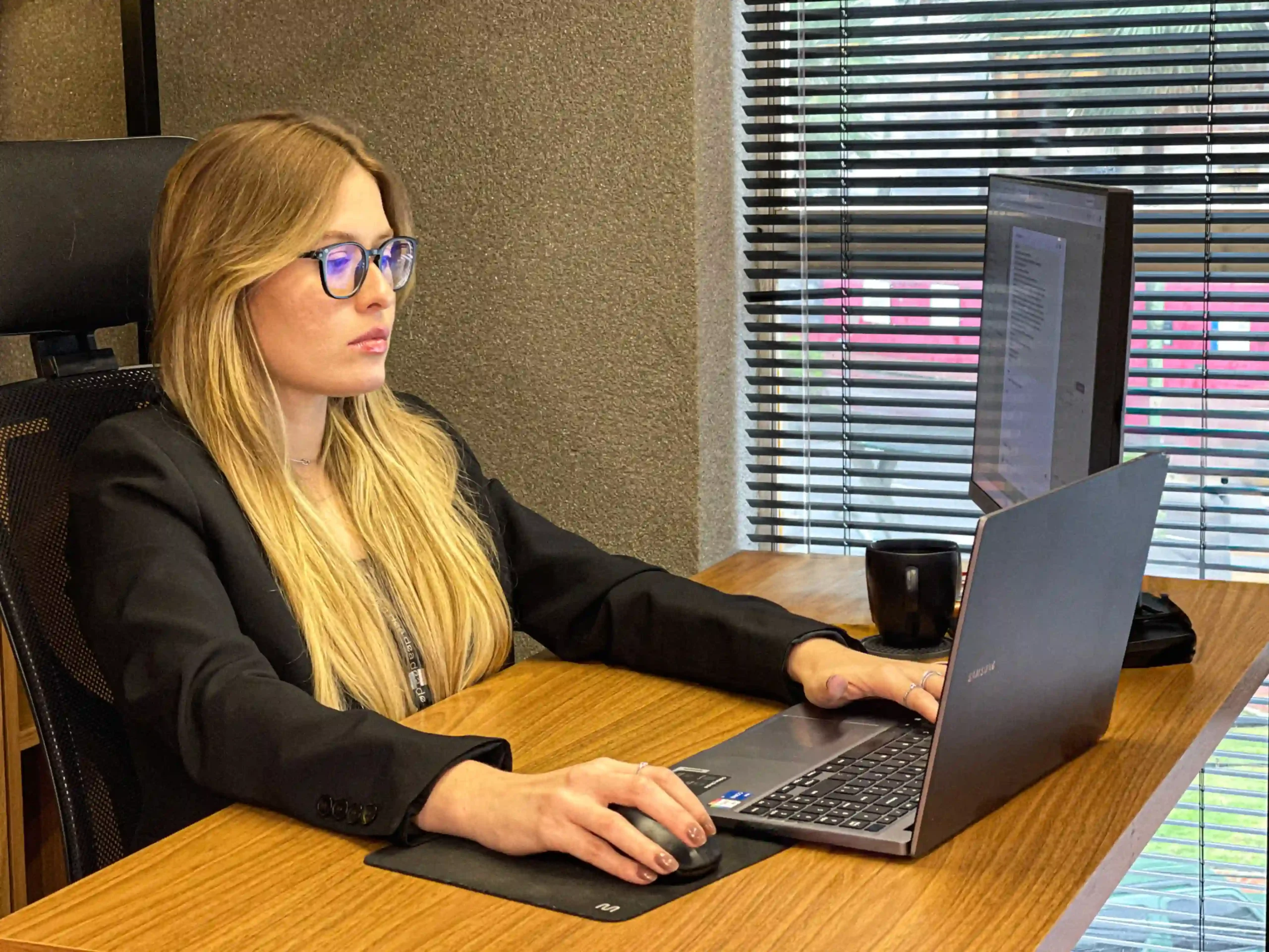 Integrante do escritório de blazer preto e óculos trabalhando em notebook e monitor