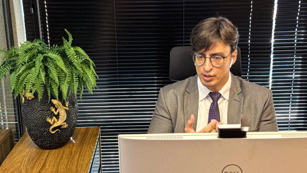 Sócio do escritório de terno cinza, gravata roxa e óculos, falando e gesticulando diante do computador