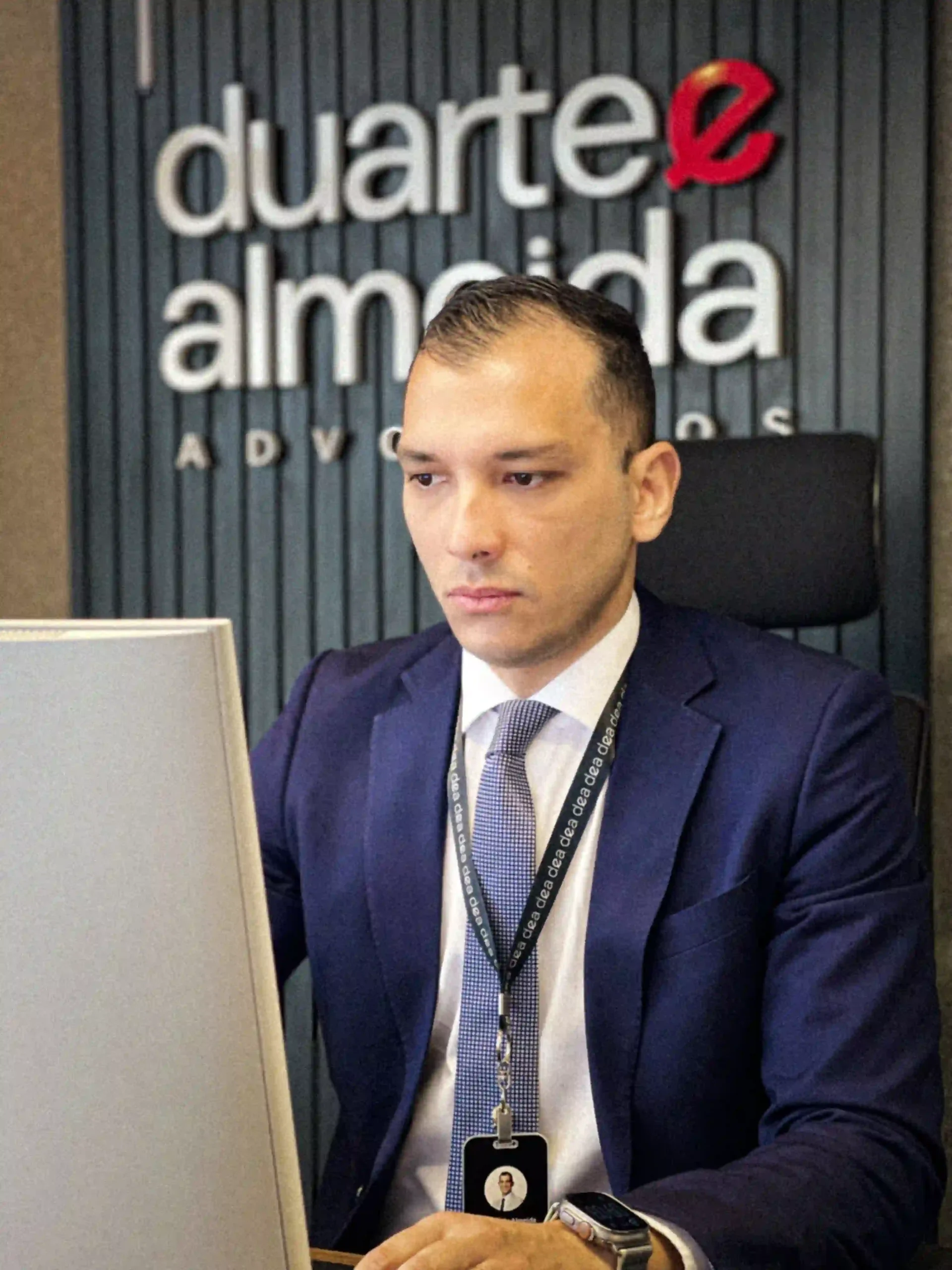 Advogado de terno azul e crachá trabalhando ao computador sob a marca do escritório